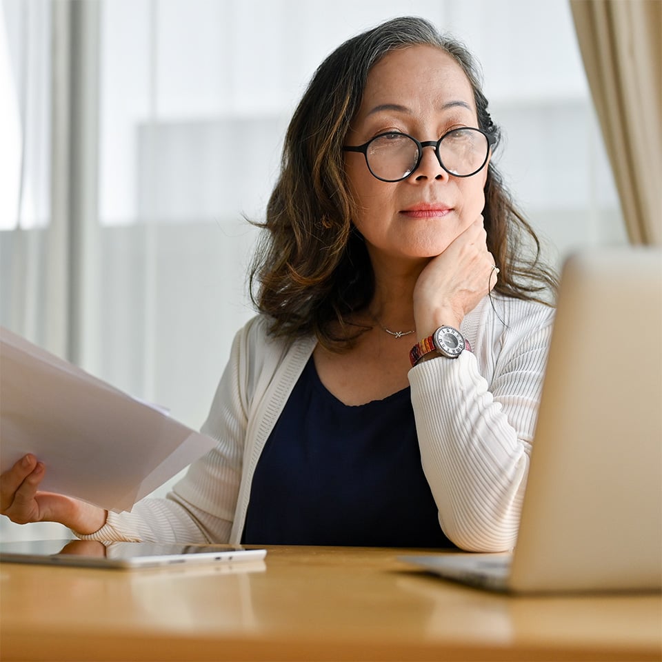 Woman wearing glasses while looking at a computer and holding papers.