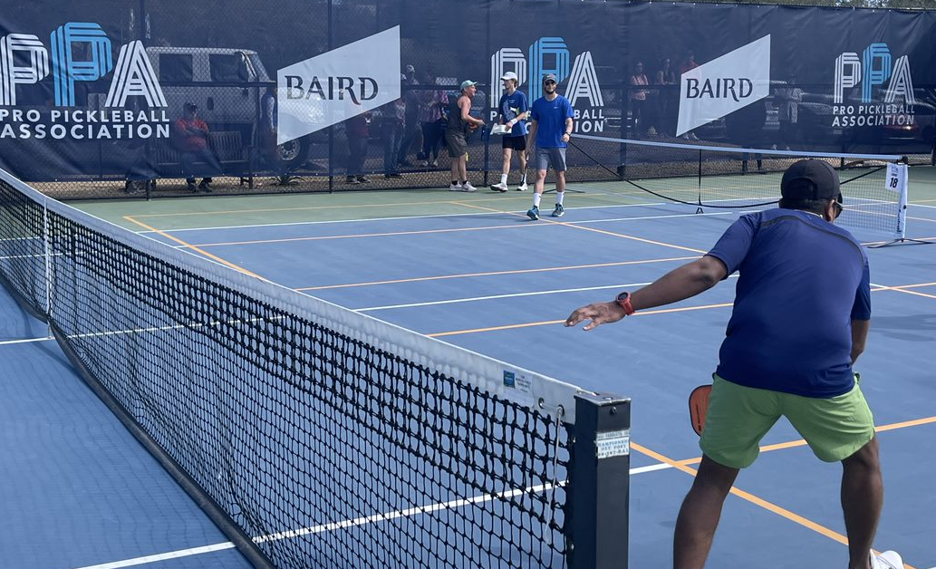People playing pickleball on a large, outdoor court. Along the perimeter of of the court is a banner printed with the the Pro Pickleball Association and Baird logos.