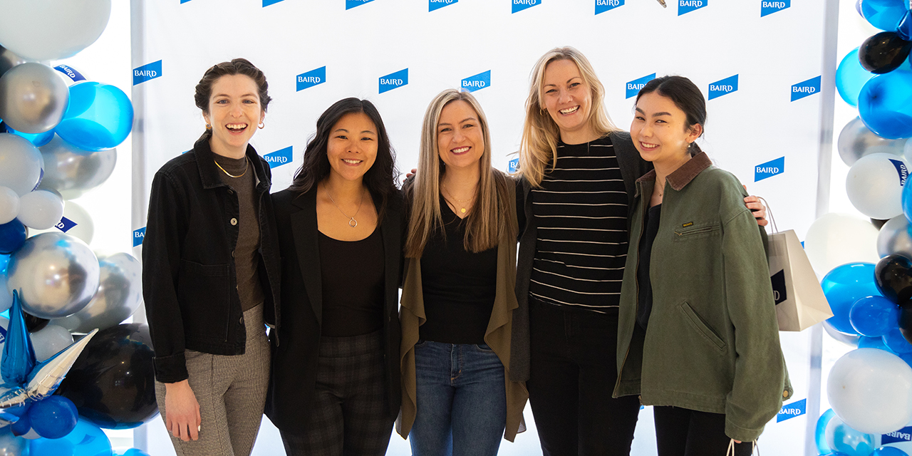 Five women stand before a Baird backdrop with blue, white, and silver balloons.