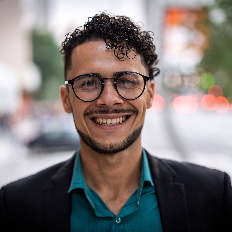 Portrait of male with round, black glasses, curly, dark brown hair and beard wearing a teal button down and black blazer with blurred street in the background.