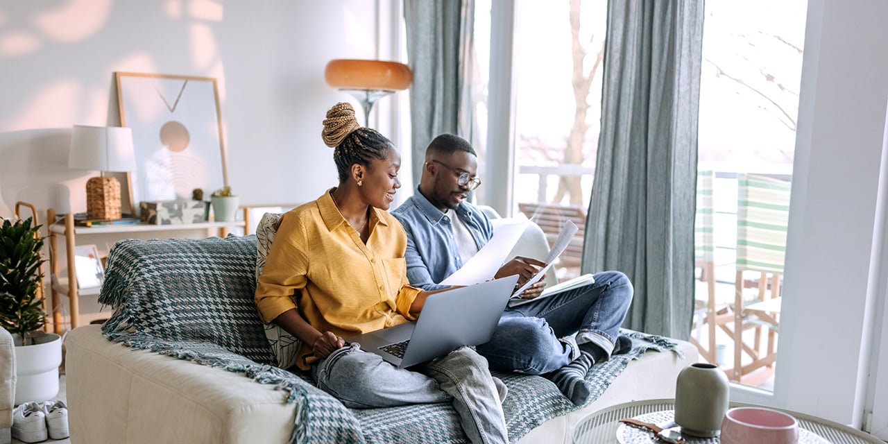 Two people sit on a couch using laptops and reviewing financial documents in a bright living room.