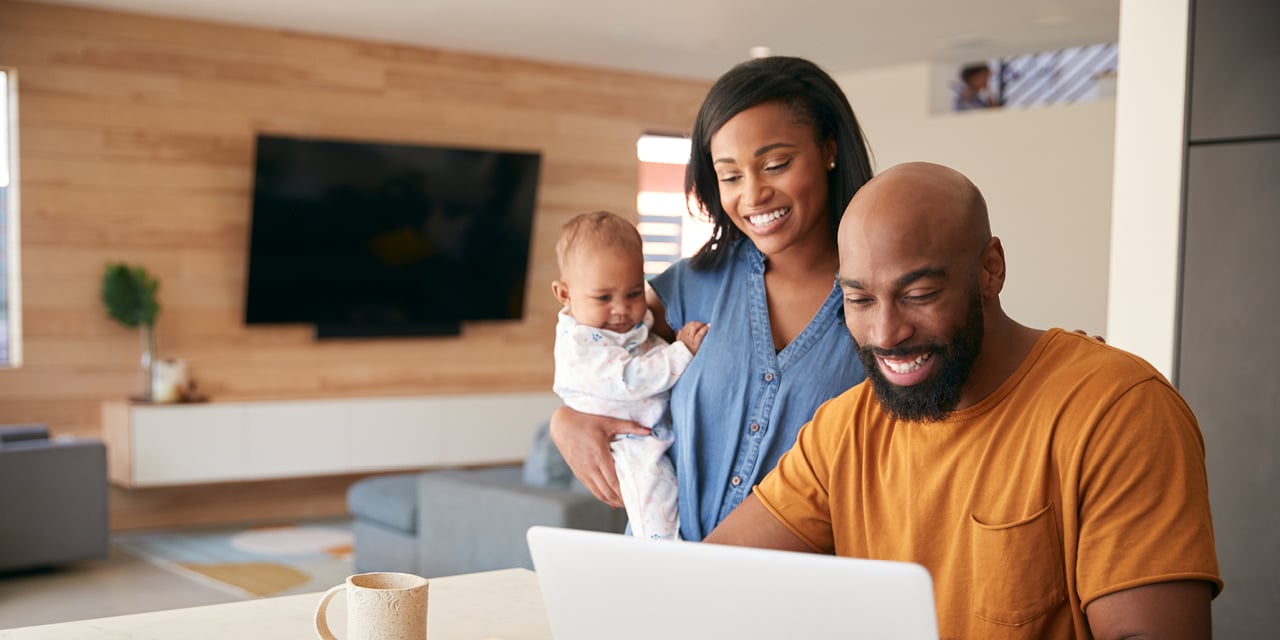 Family at home as one adult uses a laptop while another holds a baby, representing secure online financial management.