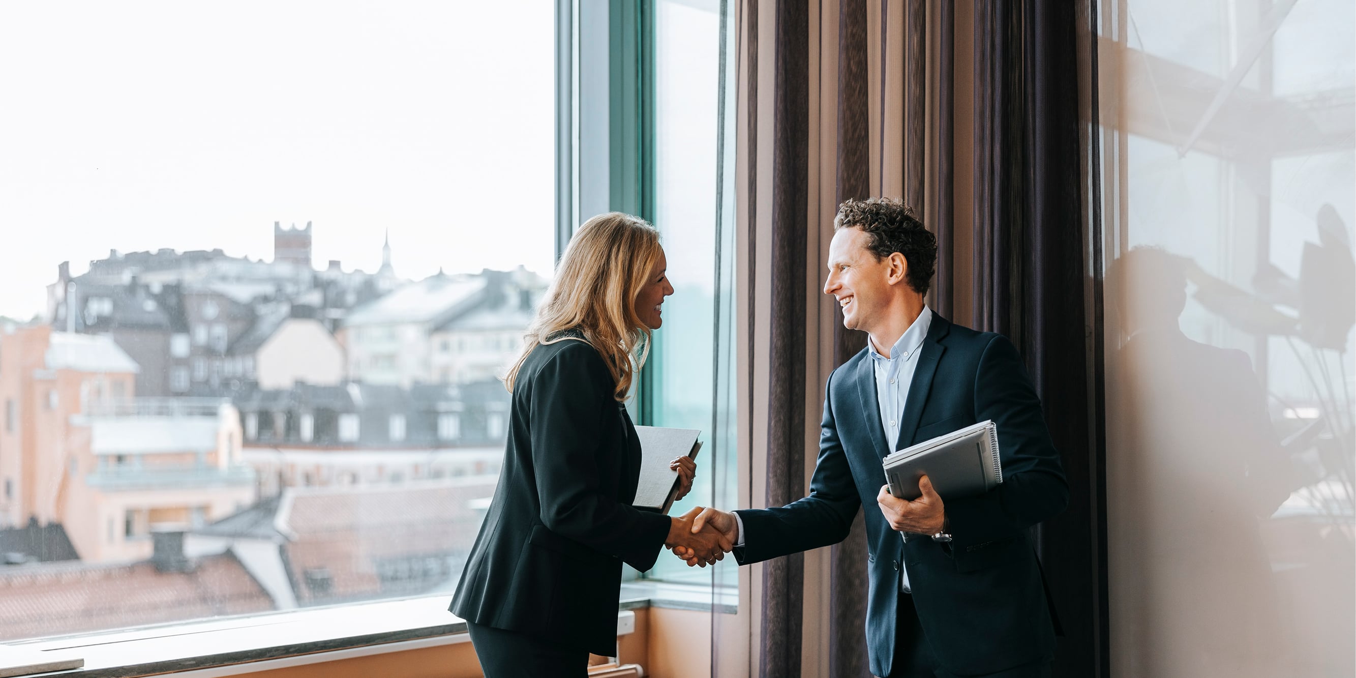 Two professionals in business attire shake hands in an office meeting room overlooking a city skyline.
