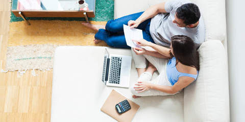 A couple sitting on a couch, reviewing a document together in front of a laptop, calculator, and other files.