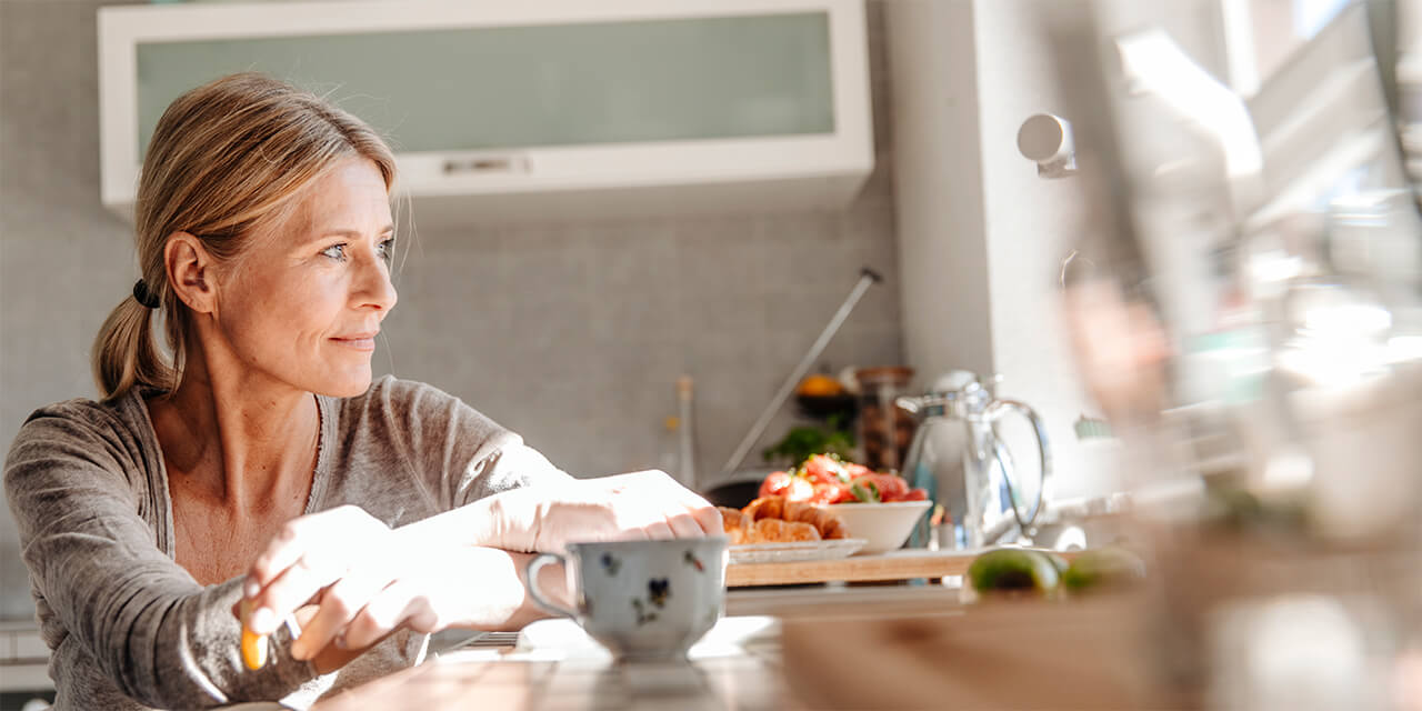 Woman eating breakfast and looking out window in a kitchen setting.
