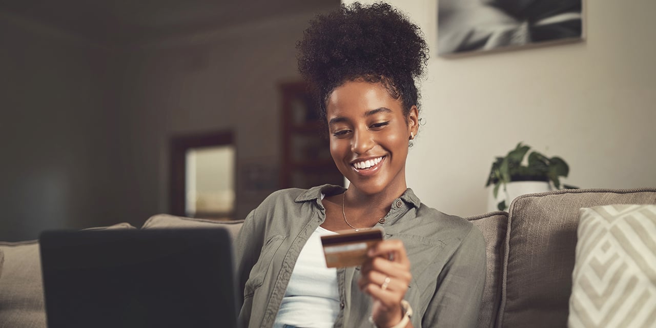 Person on couch holding credit card and using laptop, with plant and wall art in background.