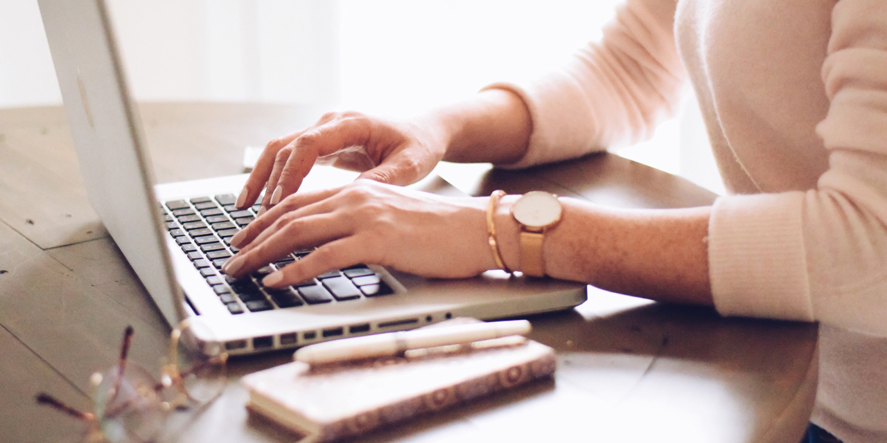 Woman typing on keyboard of a laptop computer with mobile phone, a pen and glasses on the table next to her.