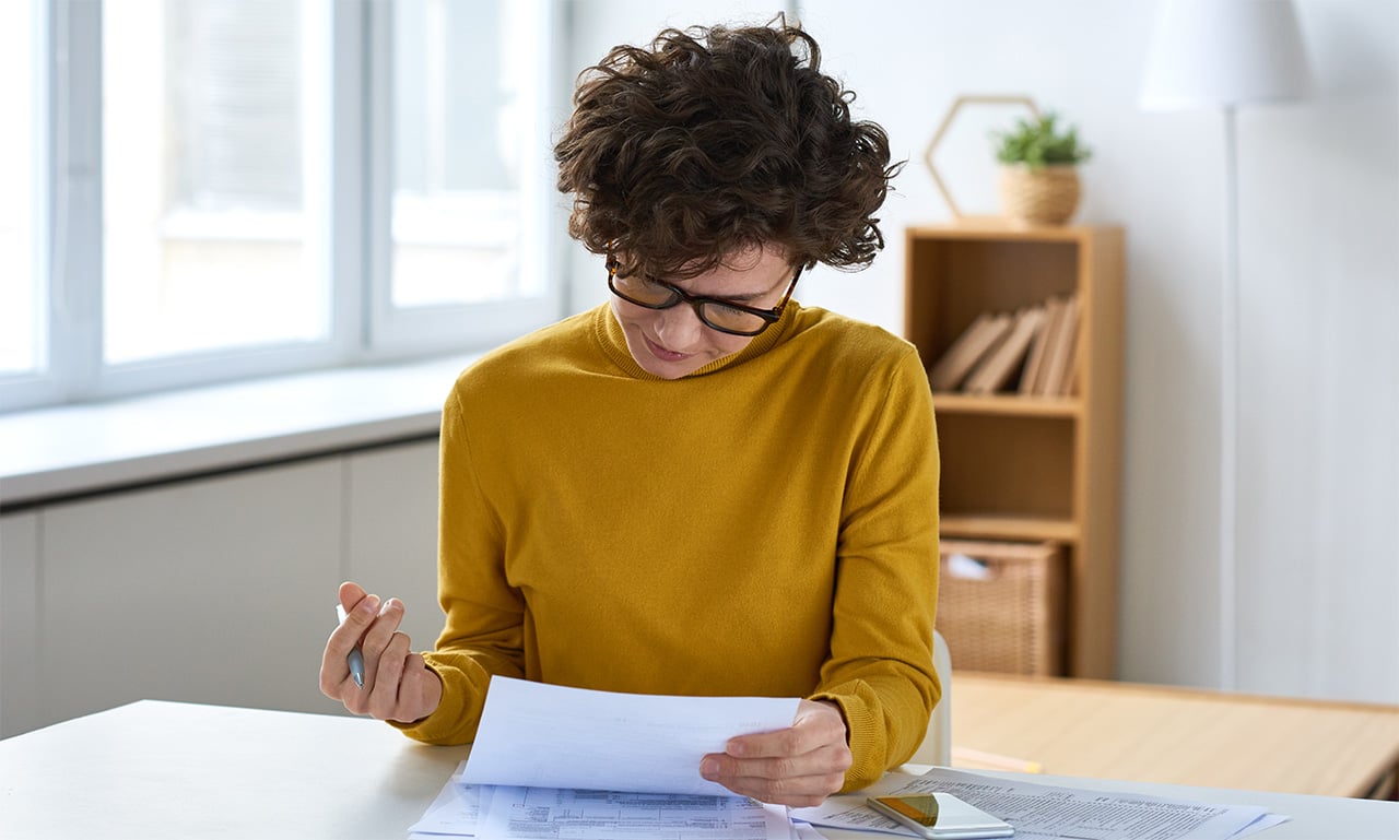 Person reviewing financial documents at a desk with forms, notes, and a calculator in a bright office.