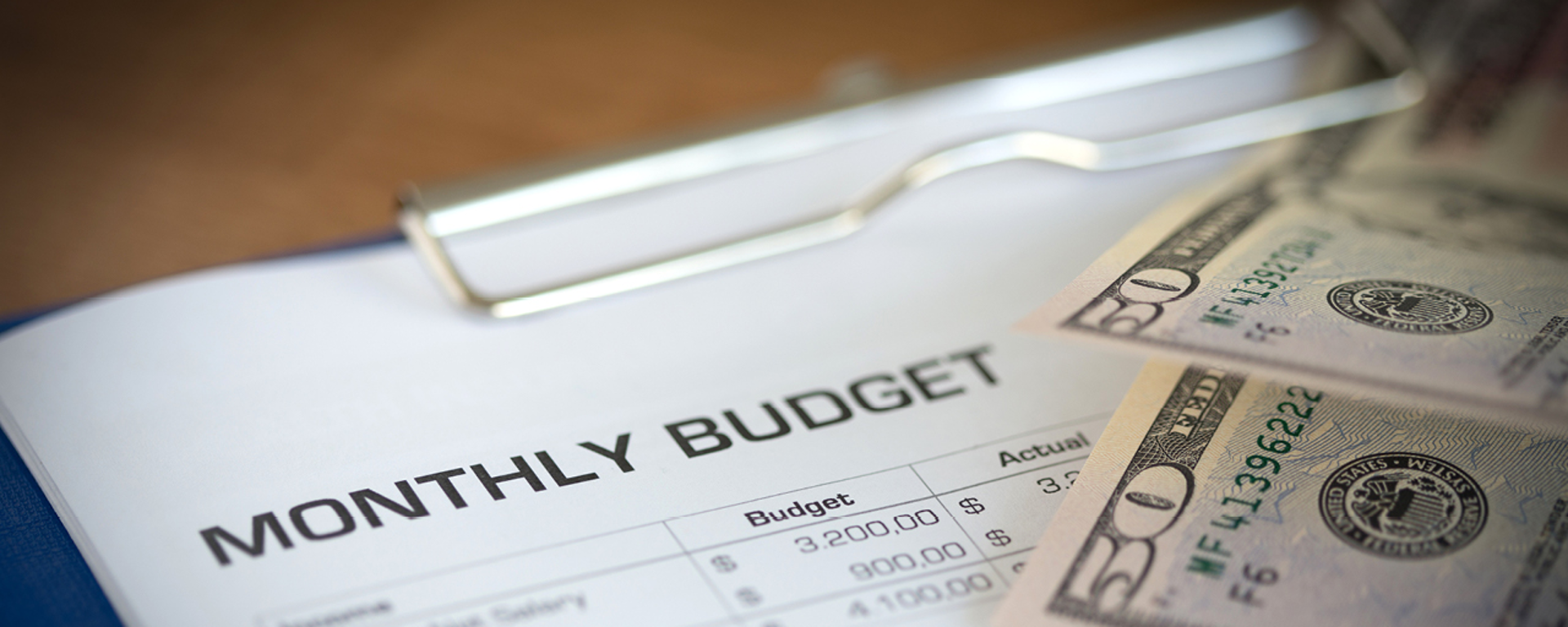 Clipboard holding a printout of a monthly budget accompanied by two $50 bills