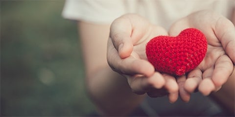 Cupped hands holding up a small, knitted red heart.