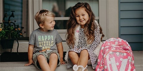 Blonde haired children sitting on front porch with backpacks.