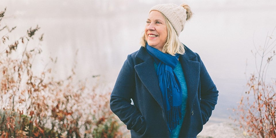 Older woman, dressed in winter clothes, smiling and looking off into distance while standing by lake.
