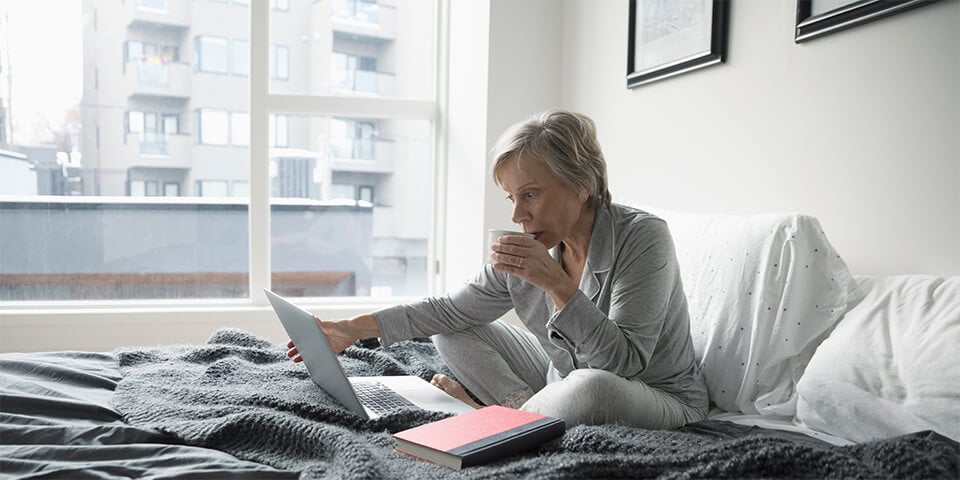 Grey-haired woman, sitting in bed and sipping coffee as she looks at laptop screen.