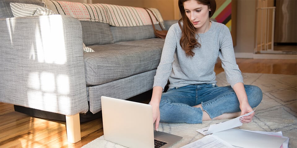 Young woman sitting on living room floor with paperwork and laptop.