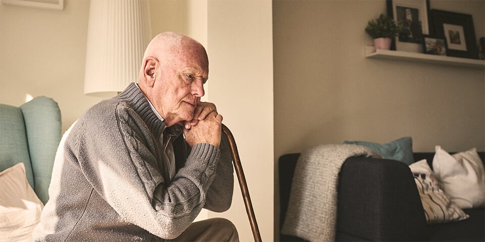 Elderly man sitting, leaning on cane in deep thought.