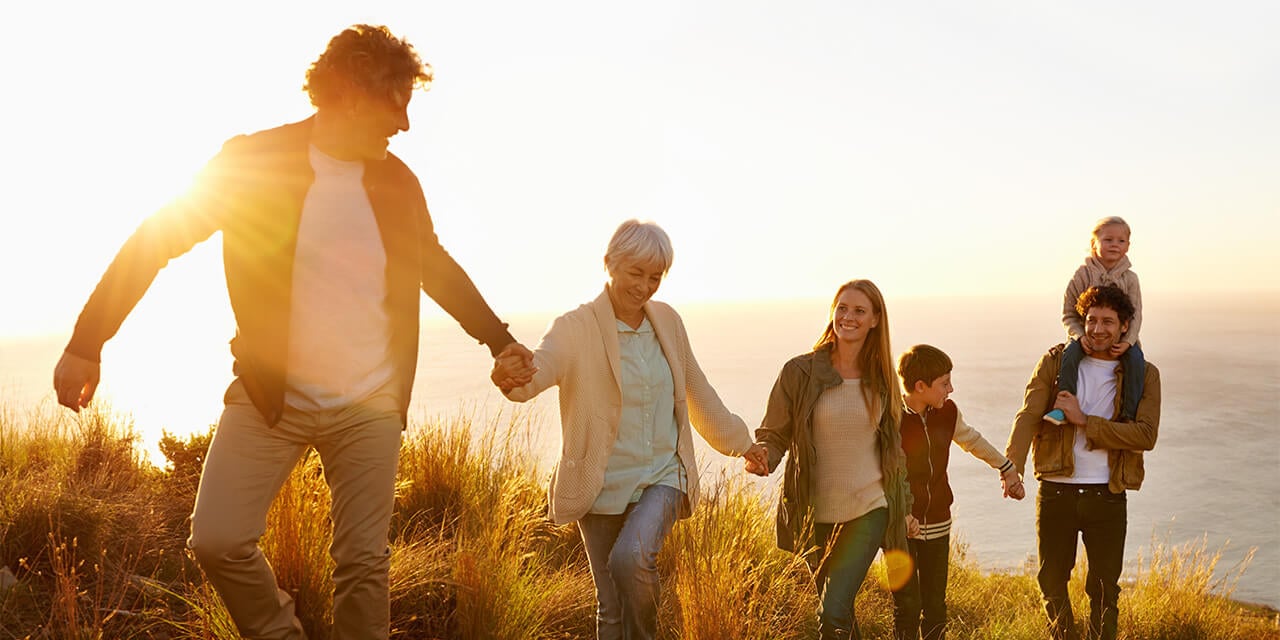 A multi-generational family of six holds hands as they walk up a grassy lakeshore