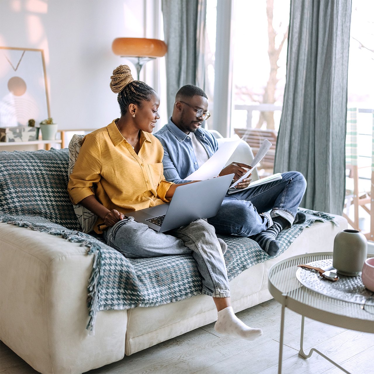 Two people sit on a couch using laptops and reviewing financial documents in a bright living room