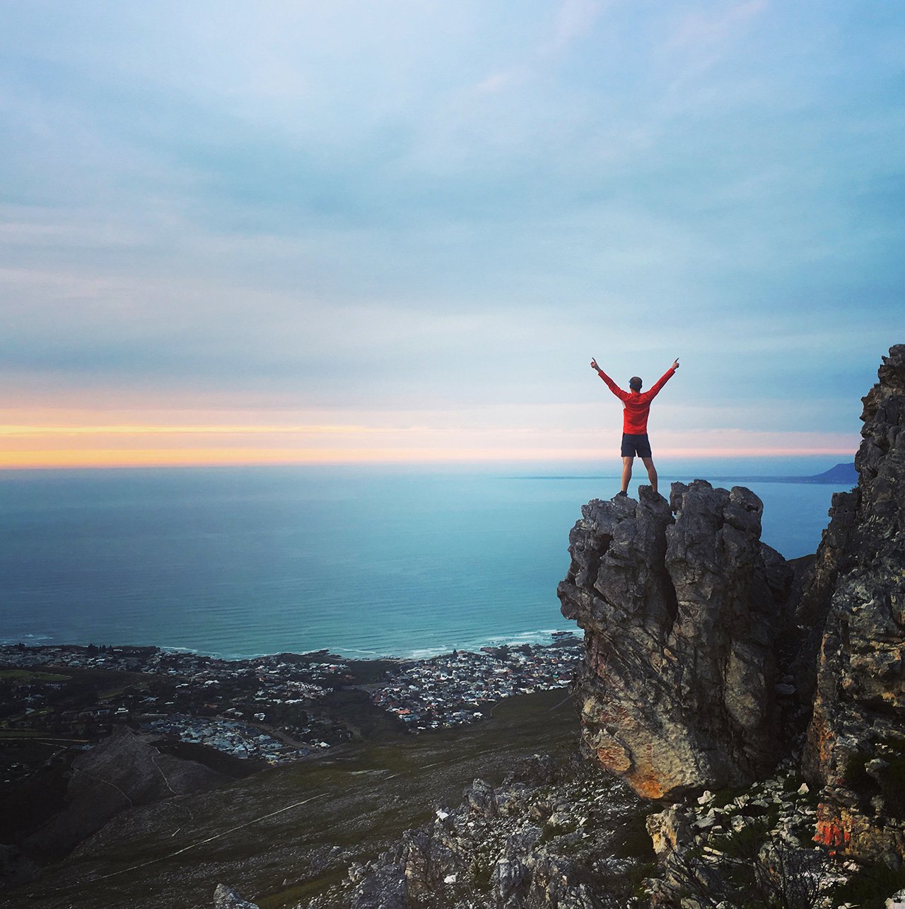 Person posing victoriously on a cliff overlooking the ocean at sunrise