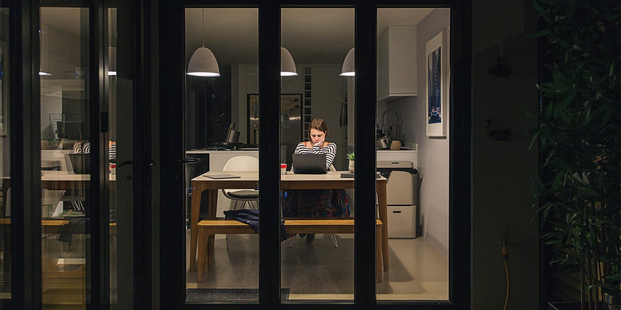 View into the window of a house at night where a woman looks concerned as she looks at a laptop