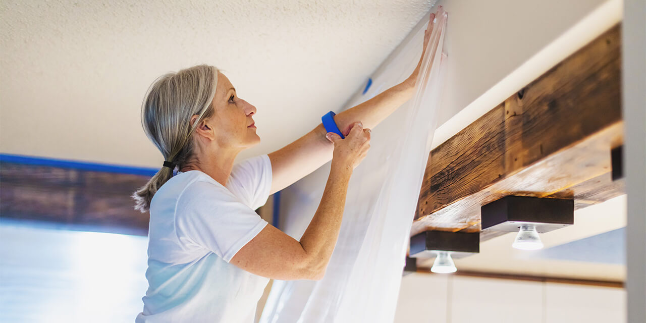 A woman standing on a ladder hanging a drop cloth with painters tape